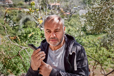 Mature bearded man sitting in the garden and talking on the phone on the speakerphone.