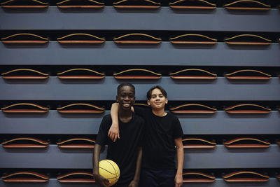 Happy teenage boy with basketball standing by male friend at sports court