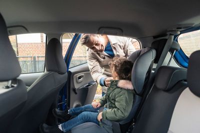 Smiling father fastening seat belt of daughter while standing by car