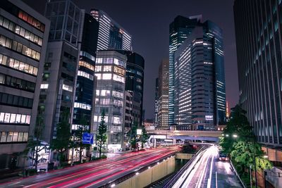 Light trails on city street by buildings against sky at night