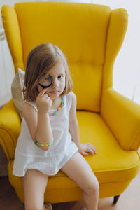 Blonde girl with a magnifying glass in her eye sitting on a yellow armchair