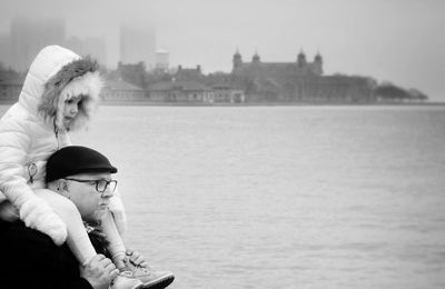 Father piggybacking daughter by river against clear sky