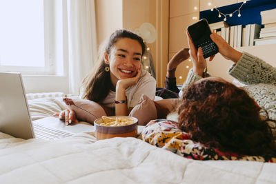 Smiling young woman using laptop while friend text messaging through smart phone on bed