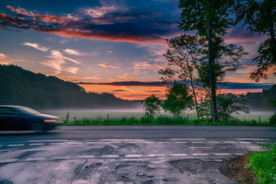 Road by trees against sky during sunset