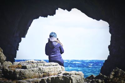 Rear view of woman sitting on rock at beach seen through cave