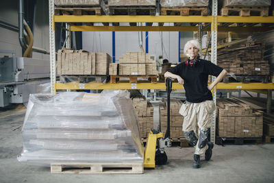 Full length of confident worker standing by stacked planks on pallet jack at industry