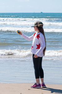 Girl taking selfie through smart phone while standing on sea shore at beach