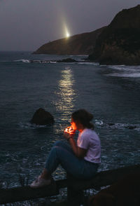 From above side view of casual pensive young female smoking cigarette while sitting on fence on rocky sea coast in summer evening in town of lekeitio in spain with beacon light in background
