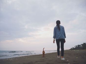 Rear view of woman walking with dog at beach against cloudy sky