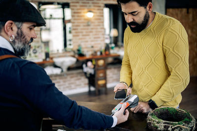 Ethnic male client making contactless paying with smartphone on pos terminal in modern grooming salon