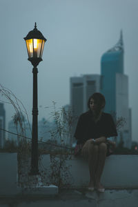 Man sitting on street light against sky in city