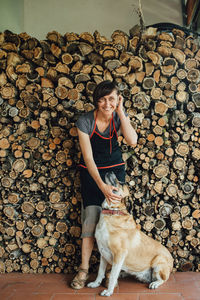 Smiling middle age woman in apron with dog at pile of sawn logs