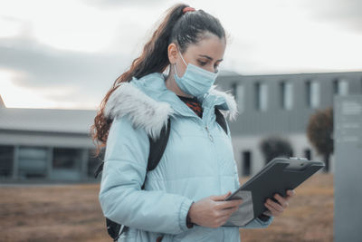 Young woman wearing mask holding clipboard outdoors