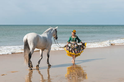 Latin female in ruffled traditional flamenco costume walking with gray stallion on sandy sea beach while looking away