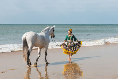 Latin female in ruffled traditional flamenco costume walking with gray stallion on sandy sea beach while looking away