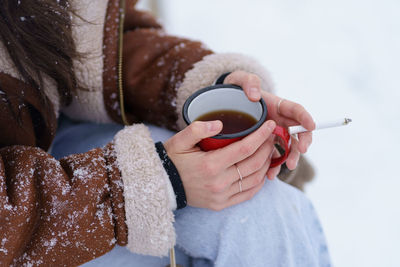 Woman drink hot tea from metal mug and smoke cigarette outdoors during cold winter snowy day