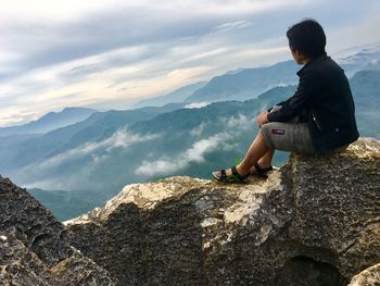 Full length of hiker sitting on rock by mountains against cloudy sky