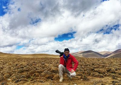 Portrait of man holding camera while crouching on field against sky