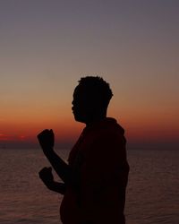 Silhouette teenage boy standing by sea against sky during sunset