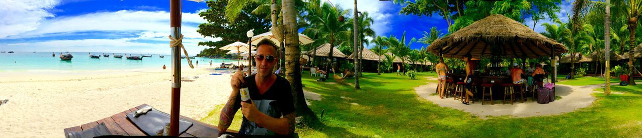 beach, palm tree, sky, tree, sand, water, sea, vacations, shore, sunlight, thatched roof, beach umbrella, lounge chair, built structure, panoramic, parasol, leisure activity, shadow, building exterior