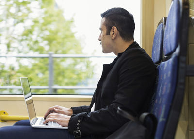 Side view of young man using laptop while looking through tram window