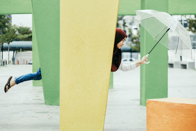 Teenage girl holding umbrella while standing behind column