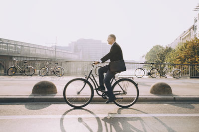 Side view of businessman riding bicycle on road in city against sky