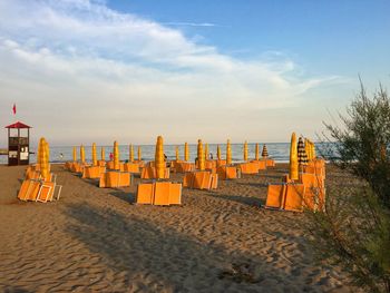Closed parasols and chairs at sandy beach against sky during sunset