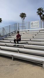 Man sitting on bench by railing against sky