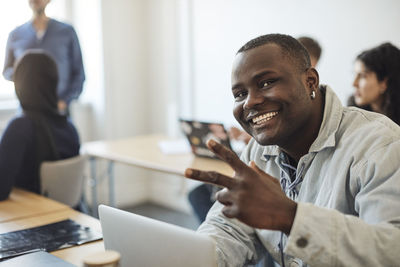 Portrait of happy male student showing peace sign while sitting in classroom