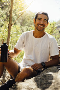 Happy male hiker holding water bottle while sitting on rock against trees in forest