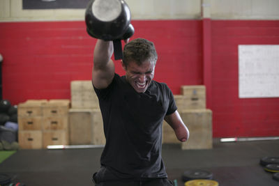 Aggressive male adaptive athlete lifting kettlebell while standing against wall in gym