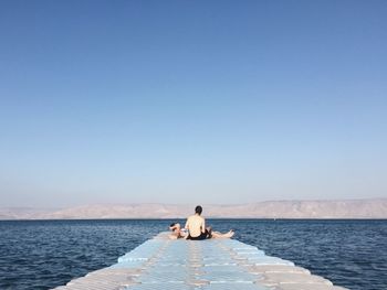 Friends resting on pier over lake against clear blue sky