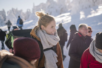 Smiling young woman wearing warm clothing while looking away by people in winter