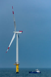 Wind turbine by ship in sea against clear blue sky