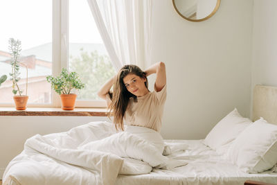 A young brunette enjoying awakening sits on a bed on bed linen near a window in a cozy apartment.