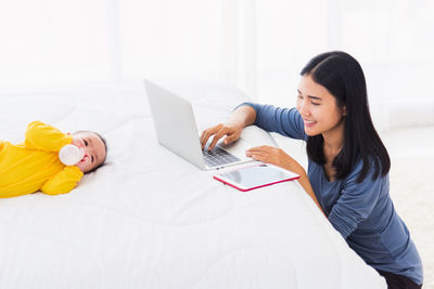 Woman using laptop while baby girl lying on bed