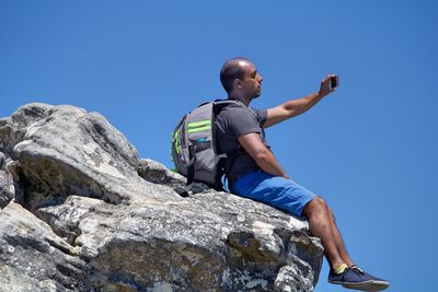 Low angle view of man taking selfie sitting on rock against sky