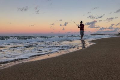 Man fishing in sea against sky during sunset