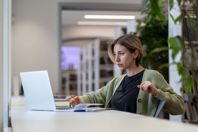 Focused pensive female university teacher preparing for online lecture on laptop in modern library