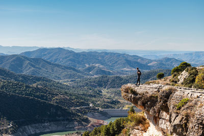 Young man with backpack standing next to cliff observing the landscape while taking a break