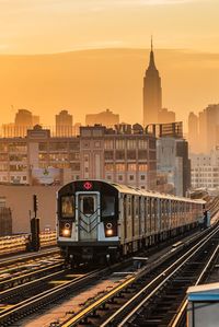 Train on railroad tracks by buildings in city against sky during sunset