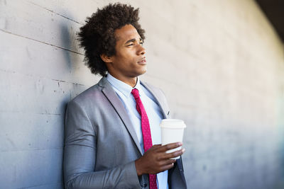 Thoughtful businessman drinking coffee while standing against wall