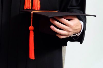 Midsection of man wearing graduation gown while holding mortarboard against white background