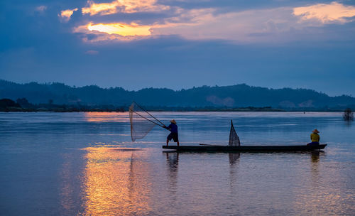 In the early morning before sunrise, an asian fisherman on a wooden boat casts a net for 