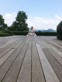 Portrait of woman sitting on wooden boardwalk against sky