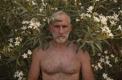 Portrait of shirtless adult man standing against plants with white flowers 