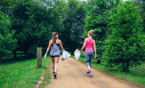 Rear view of friends walking on footpath while holding plastic bags against trees