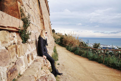 Side view of woman sitting on stone wall by sea against cloudy sky