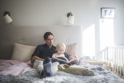 Father reading picture book with toddler son while sitting on bed at home